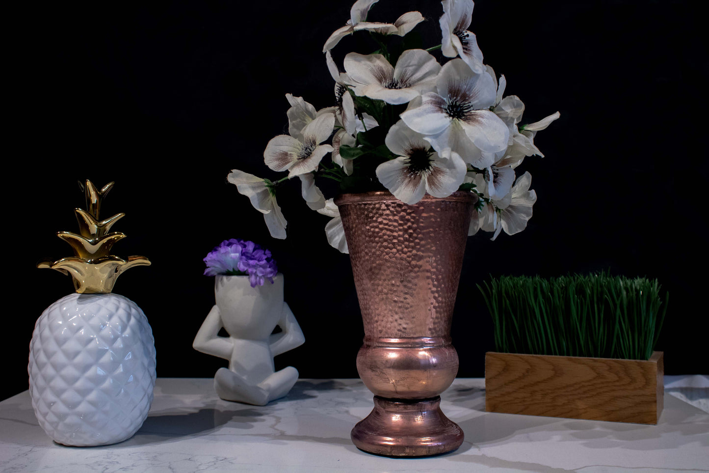 Un conjunto de cuatro adornos: una piña blanca con dorado, una maceta de "no oigo", un jarrón cobrizo en forma de cáliz alargado con flores blancas y un adorno rectangular de madera con pasto verde