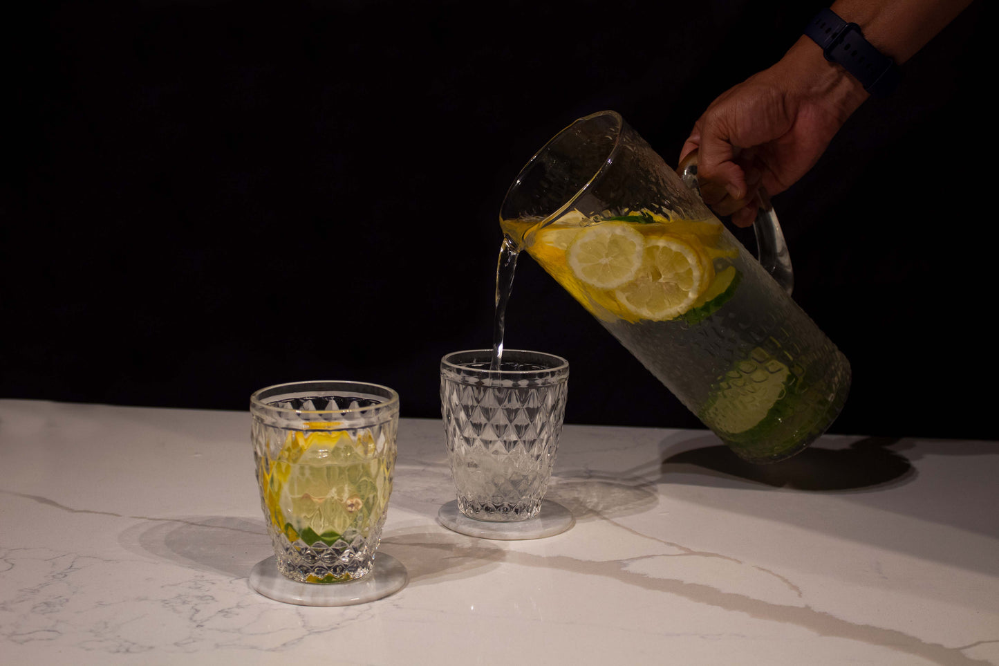 una mano sirviendo agua de una jarra con rodajas de limones y cristal transparente con textura de gotas sobre vasos de cristal transparente