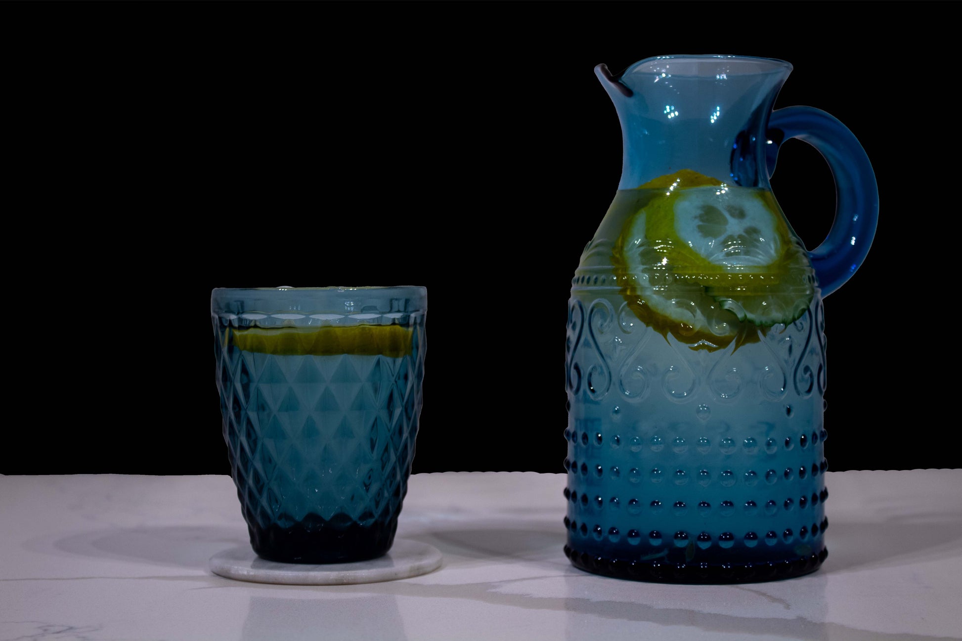 un vaso y jarra azules con estilo de vidrio soplado y diseño de grecas y diamante llenos con agua y rodajas de limón, 