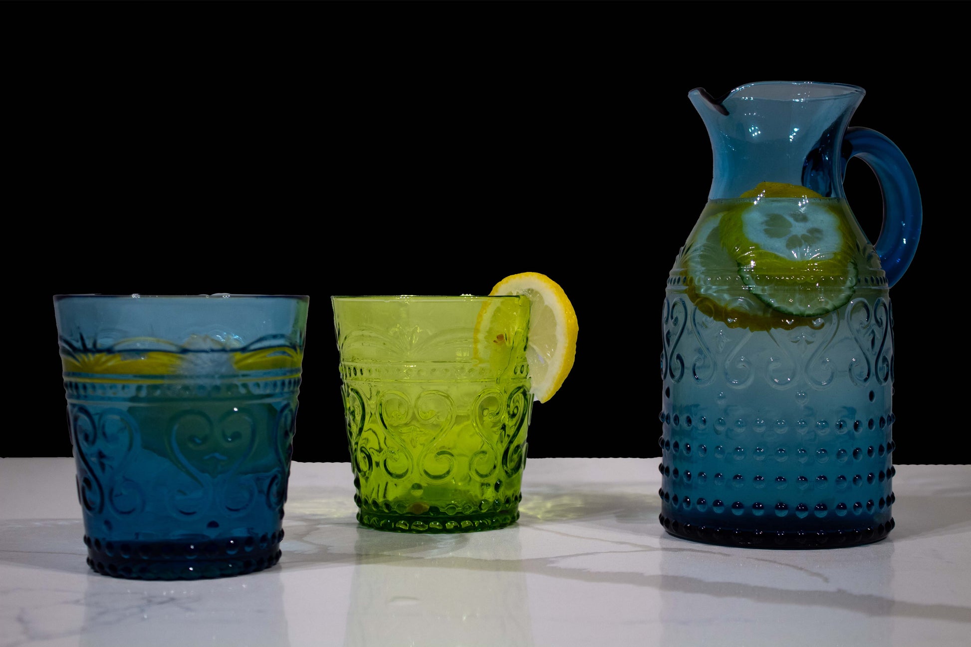 Dos vasos, uno verde y otro azul junto a una jarra azul y asa redondeada llenos con agua y rodajas de limón, los tres con diseño de grecas y estilo de vidrio soplado con fondo negro