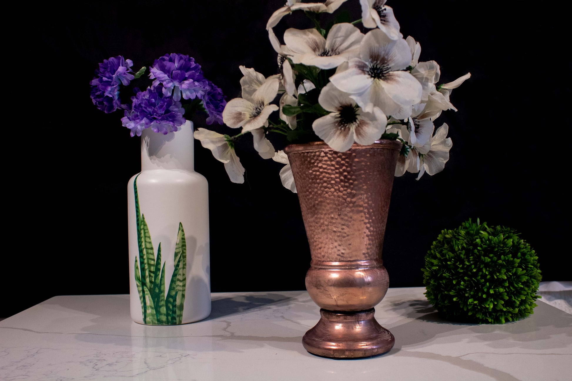 Dos jarrones floreros de cerámica, uno blanco con flores moradas y diseño de hojas verdes y con flores azules y el otro color cobrizo en forma de cáliz alargado con flores blancas, junto una bola de follaje