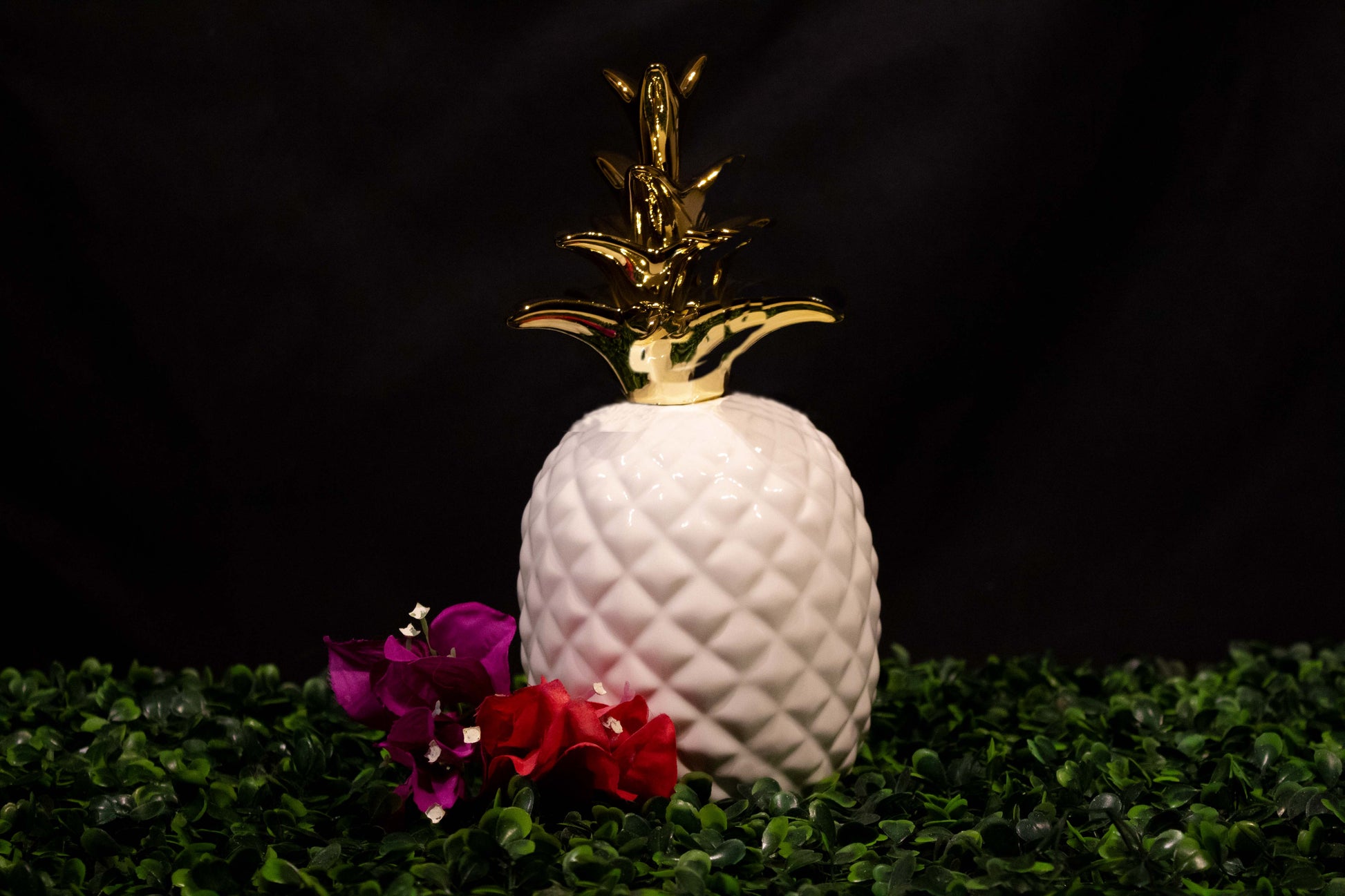 Piña de ornato de cerámica de color blanco brillante con rama dorada sobre un follaje verde junto a dos flores de bugambilia