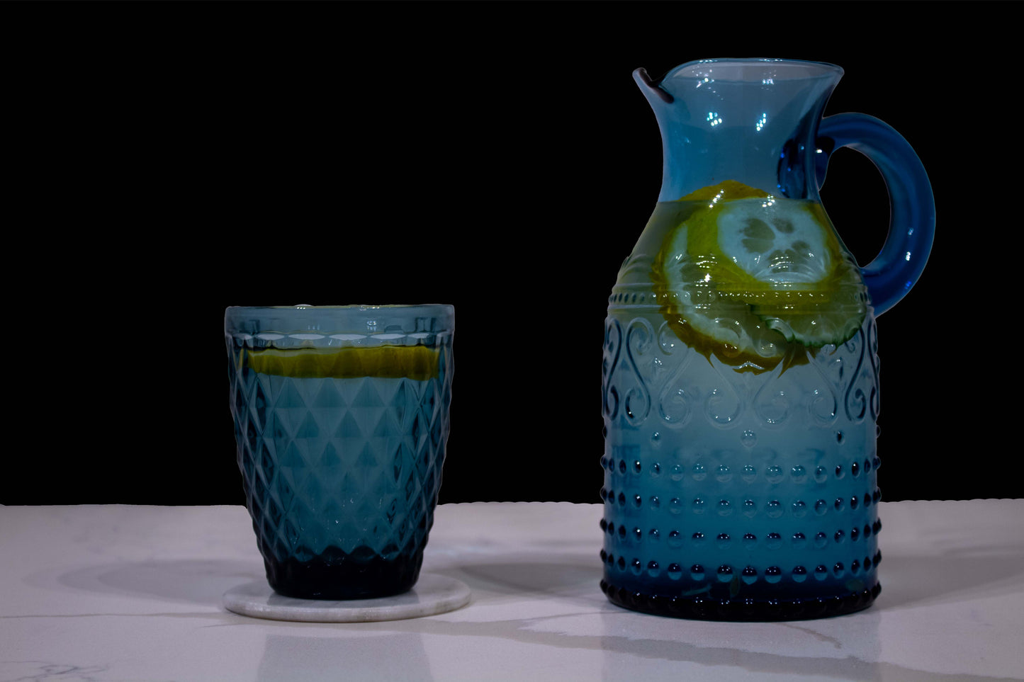 Vaso de cristal azul con diseño en forma de diamante junto a una jarra azul de cristal con diseño en grecas, llenos de agua con rodajas de limón